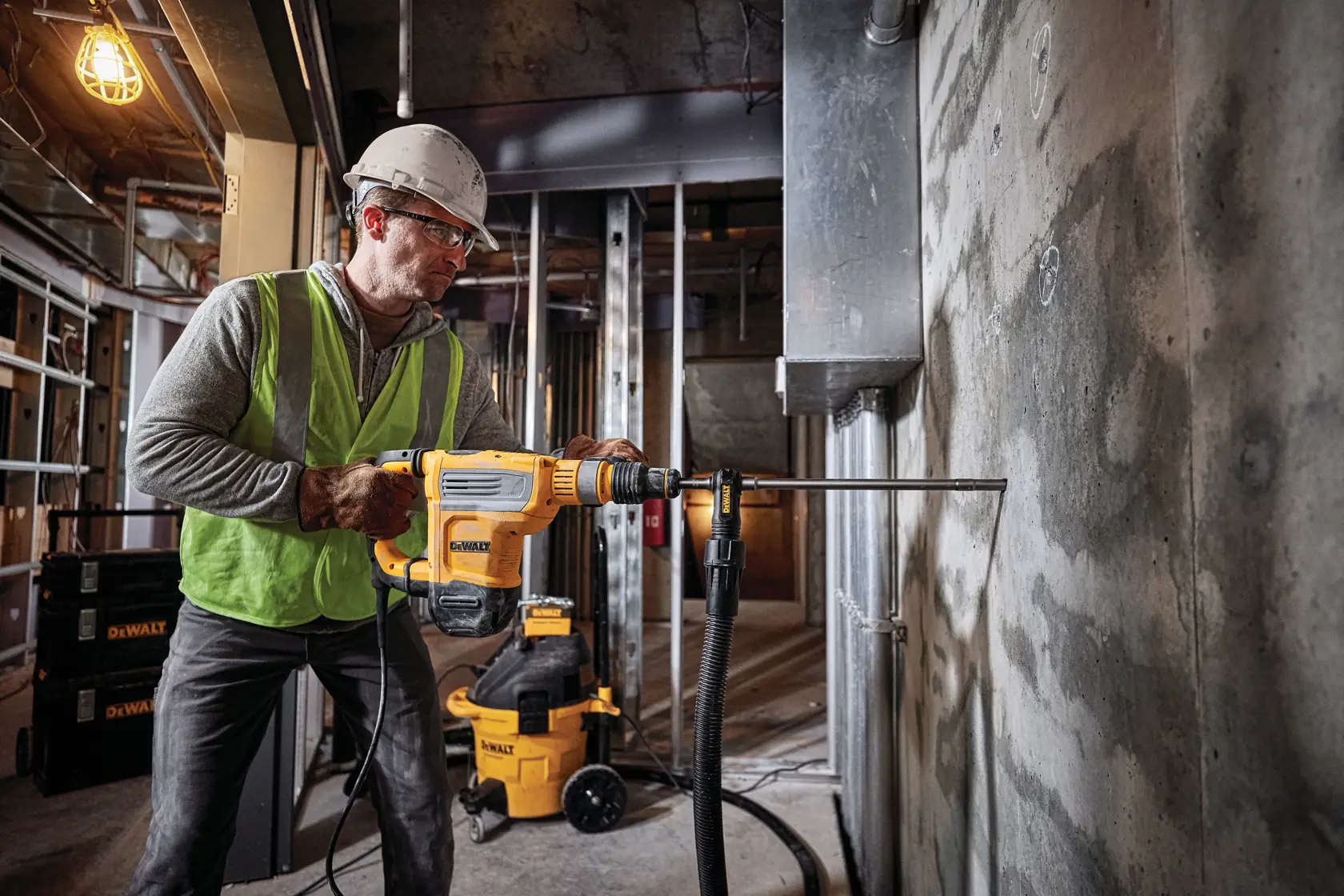 A construction worker wearing a safety helmet and vest operates a yellow DEWALT D25614K demolition hammer drill with dust extraction on a brick wall.