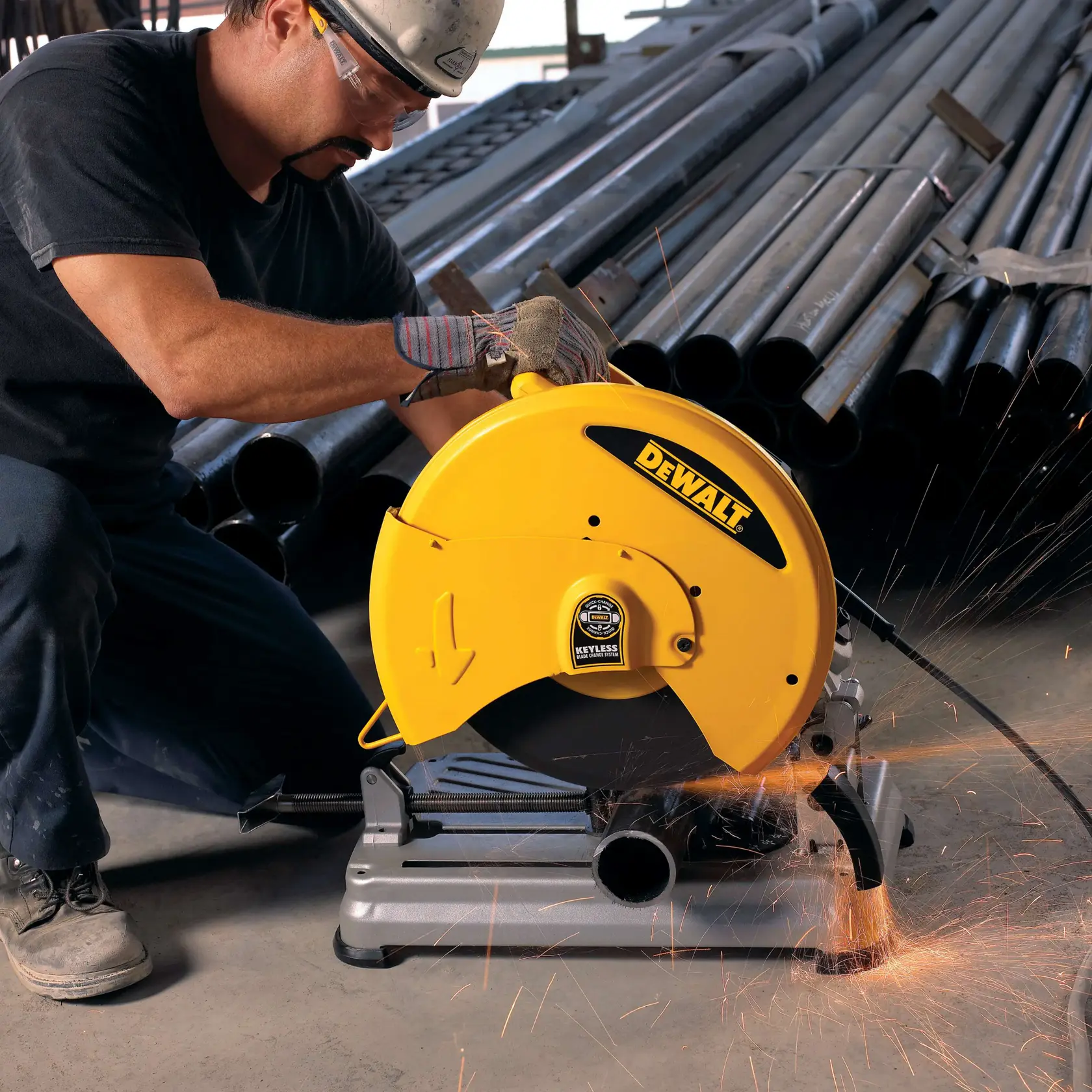 A person wearing work gloves is using a DEWALT D28715 yellow metal cutting chop saw in an industrial setting. Sparks are flying from the saw as it cuts a metal pipe, with stacks of metal tubes visible in the background.