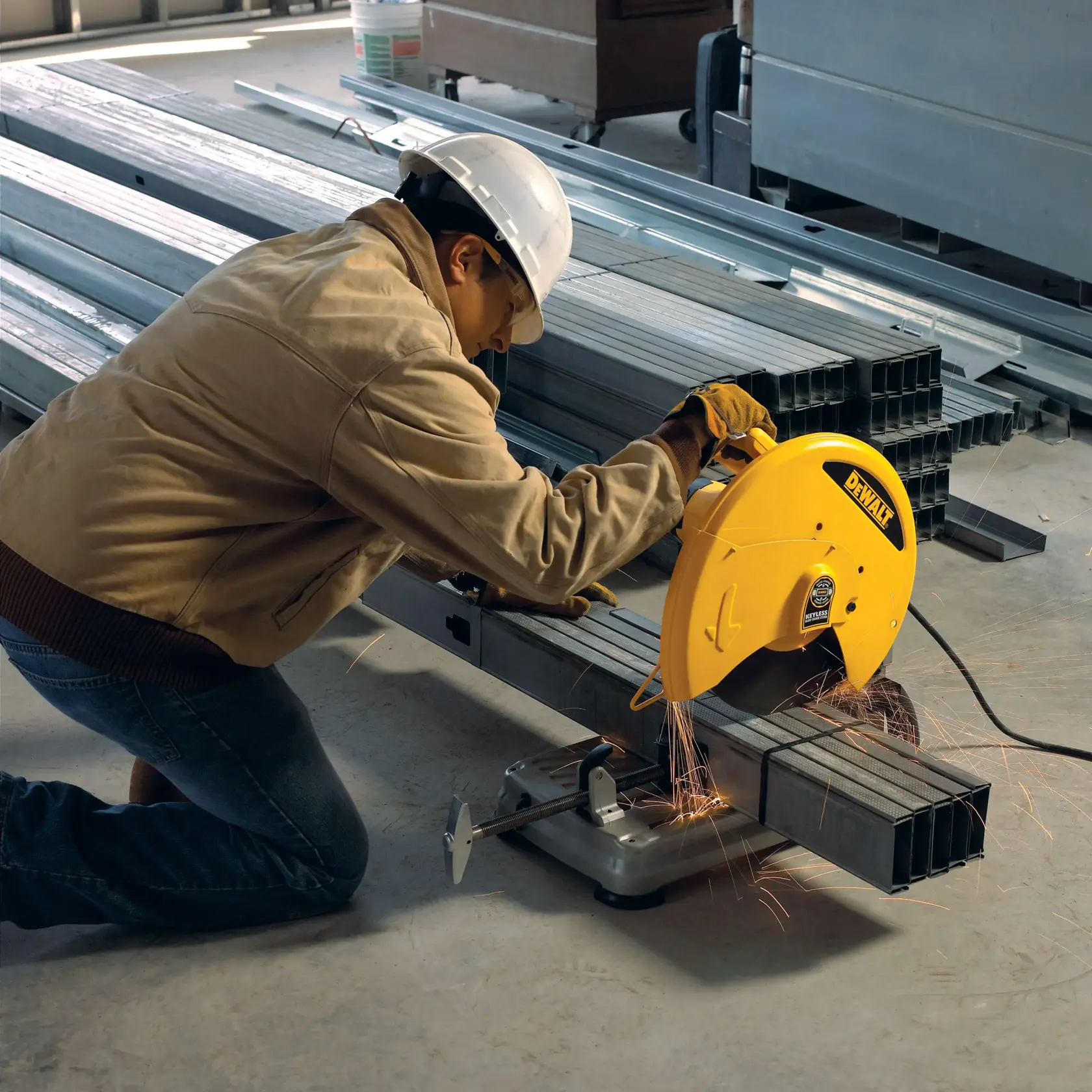 A person wearing protective gear, including a hard hat and gloves, is using a DEWALT D28715 metal cutting saw to cut steel rectangular tubes in an industrial setting. Sparks are visible, indicating active cutting.
