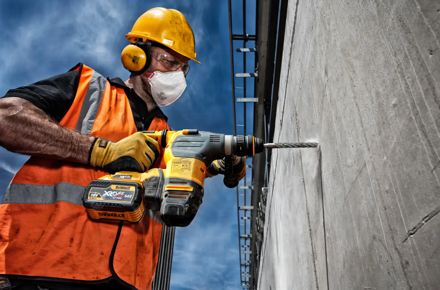 A construction worker in a safety helmet and high-visibility vest is using a DEWALT cordless rotary hammer drill to drill into a concrete wall.