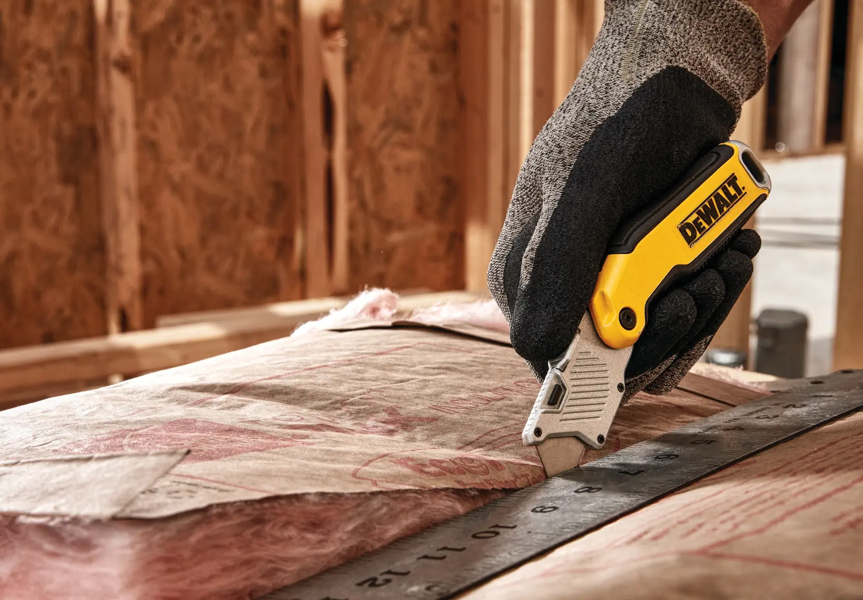 A gloved hand using a yellow and silver DEWALT utility knife to cut insulation material along a metal ruler at a construction site.