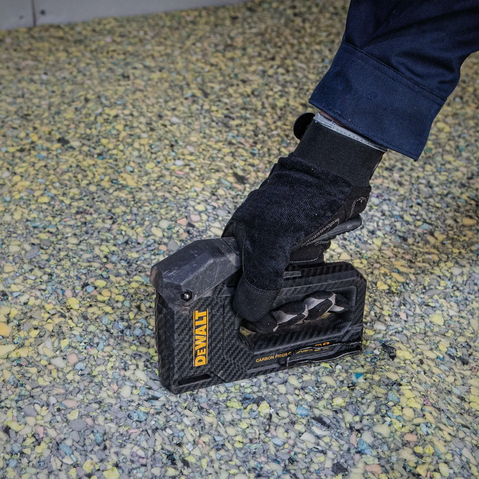 A person wearing a black glove is using a DEWALT carbon fiber hand tool, likely a staple gun, on a textured surface with yellow and multicolored speckles.