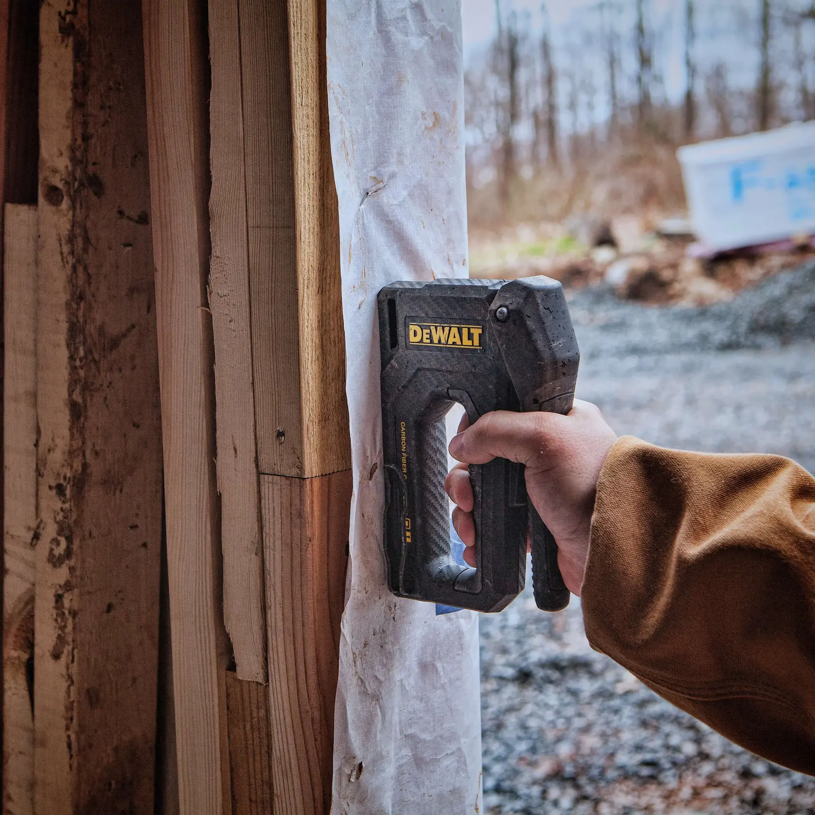 A person wearing black gloves using a DEWALT staple gun to secure insulation material to a wooden wall.